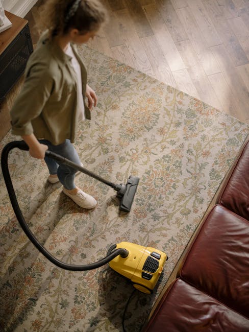 A person in full protective gear, including a white coverall, face mask, and gloves, is engaged in deep cleaning a plush, beige carpet in a modern living room. They are using a professional steam or vapor cleaning machine, with a black hose connected to the device, emitting steam onto the rug's surface. The room features a wooden shelving unit with decorative items, a tall mirror, and potted plants, illuminated by natural light. The clean and well-maintained space highlights the importance of surface cleaning and sanitisation, as performed by Carpet Cleaning Finsbury Park, to ensure hygiene and maintain fresh, healthy indoor environments.