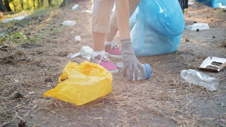 Close-up of a person's hands wearing grey gloves, collecting plastic water bottles and trash from the ground during a community clean-up event in an outdoor park area with dirt and pine needles. The p