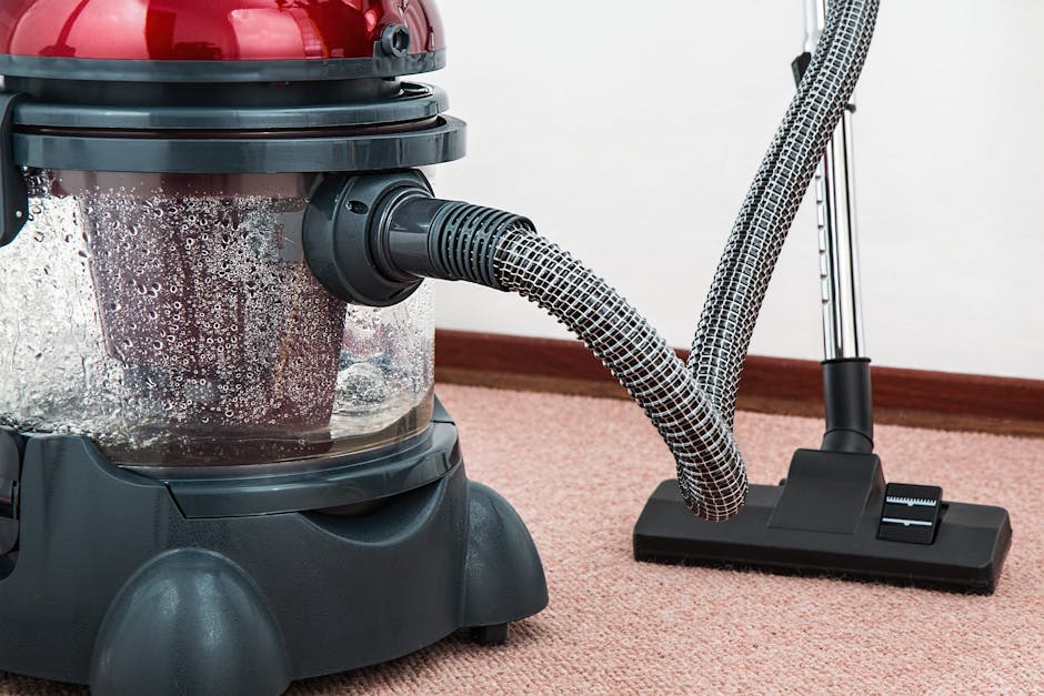 A woman with curly hair, dressed in a beige jacket, blue jeans, and white sneakers, is performing surface cleaning with a vacuum cleaner on a large patterned carpet in a living room. She is holding th