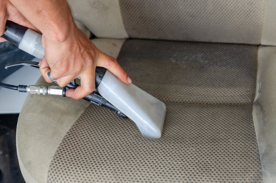 A person using a handheld upholstery cleaning machine on a beige fabric car seat or sofa cushion, which has a textured area with small perforations. The cleaning device, connected to a hose, is pressed firmly against the fabric, and the surface appears damp from the cleaning process. The surrounding area of the upholstery is clean, and the scene is well-lit, highlighting the surface being cleaned. The image exemplifies professional surface cleaning and sanitisation service offered by Carpet Cleaning Finsbury Park, focusing on deep cleaning and hygiene of upholstered furniture surfaces.